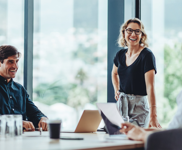 A group of four business professionals is in a meeting room with large windows overlooking a city. A woman with glasses and a black shirt stands smiling at the center. To her left, a man in a dark blue shirt is laughing, and a woman is looking at him. To her right, a person in a white shirt is turned towards her. A laptop and coffee cup are on the table.